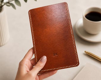 May include: A rectangular brown leather phone case with white stitching around the edges. The initials "JFK" are embossed on the lower right corner. The case is held by a hand.