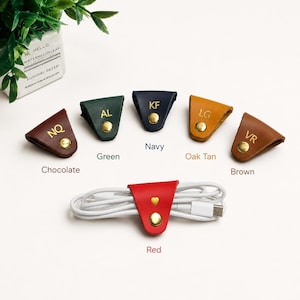 May include: A collection of six triangular leather cable organizers in chocolate, green, navy, oak tan, brown, and red. Each organizer features a gold-tone metal snap button and gold embossed initials or a heart symbol on the front surface.