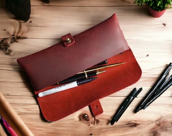 May include: A burgundy leather pencil case with a brass closure, open to reveal a variety of pens and pencils. The case is lying on a wooden surface.
