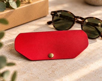 May include: A bright red, flat, hexagonal-shaped glasses case featuring a single brass-colored metal snap button closure at the center. The case is positioned horizontally, showcasing its smooth, solid-colored surface and clean, angular edges.