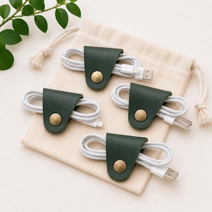 May include: Four dark green leather cable organizers with brass-colored metal snap buttons, each securing a white charging cable. The organizers are arranged on a beige cotton drawstring pouch, with a small green leafy branch visible nearby.