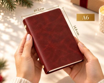 May include: A dark red A6 notebook with a smooth, textured cover and two gold-colored metal studs on the side. The notebook measures 10.5 cm in width and 14.8 cm in height as indicated by the text and dimension lines on the image.