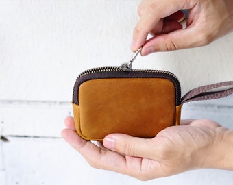May include: A small, brown leather zipped pouch with a brown strap. The pouch is being held open by a hand.