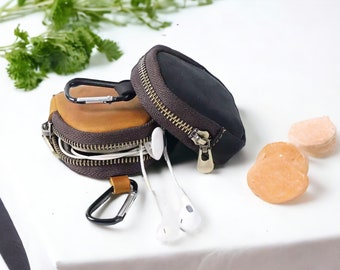 May include: Two small zippered pouches, one brown leather and one black fabric, with a silver carabiner clip attached. The pouches are open and contain white earbuds.