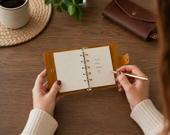 May include: A small, mustard-yellow leather ring-bound notebook with a snap closure. The open pages feature a grid pattern on the left and a list on the right with the handwritten text: Focus, Plan, Create, and a heart symbol. A gold pen is held nearby.