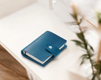 May include: A small, blue leather-bound notebook with a silver button closure. The notebook is lying on a white surface.