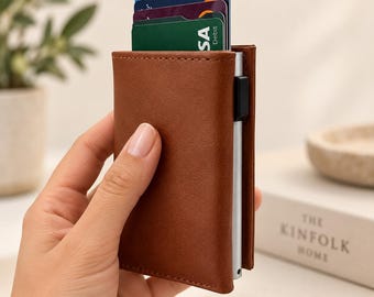 May include: A brown leather card holder wallet with a silver metal card ejector mechanism on the side. The wallet contains several credit and debit cards, including a green card with visible text, stacked vertically within the metal compartment.