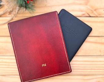 May include: A burgundy leather Kindle cover with white stitching and the name "Phil" in gold lettering. A black Kindle device with the Amazon logo is next to the cover. The items are on a wooden surface.