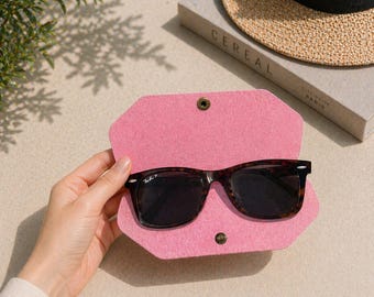 May include: Tortoiseshell sunglasses sit in an open pink felt case, secured with a snap closure. A straw hat and a book titled "Cereal" are in the background. The case is likely for protecting the glasses.