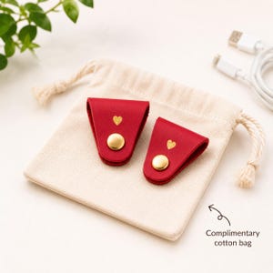 May include: Two red leather cord organisers with gold-tone snap buttons and small gold heart icons, resting on a beige cotton drawstring pouch. The pouch features the text Complimentary cotton bag with an arrow pointing toward the fabric.