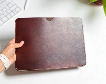 May include: A brown leather laptop sleeve with tan stitching. The rectangular case has a textured surface and a small cutout at the top. The sleeve is held by a person's hand. The text on the image says "Leather Colour: Brown" and "Thread Colour: Tan".
