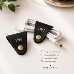 May include: Two black leather cable ties with gold-tone metal snap buttons and the embossed letters HW. One tie is folded flat, while the other secures a white charging cable. Text on the image reads: Keep your cables tidy, Personalised for you, Practical & stylish, Perfect gift idea, Small Cable Tie.
