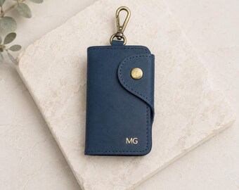 May include: A navy blue leather key case featuring a brass-toned snap button closure and a matching brass-toned swivel lobster clasp at the top. The front of the key case is embossed with the gold-colored initials MG in the bottom right corner.