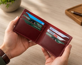 May include: A deep burgundy leather bifold wallet featuring multiple interior card slots. The wallet is open, displaying several credit cards, including those labeled Lloyds Bank and Platinum, along with a UK driving license tucked into the pockets.