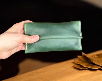 May include: A green leather wallet with white stitching is held. The wallet has a folded design and a smooth texture. The background is dark, with a wooden surface and dried leaves in the foreground. The wallet is approximately 15 cm long.
