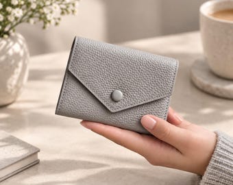 May include: A small, light gray wallet with a pebbled leather texture and a triangular flap closure. The wallet features a single matching gray snap button centered on the flap, providing a secure and compact design for everyday use.