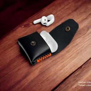 Può includere: Custodia in pelle nera per AirPods con cuciture arancioni, aperta per rivelare una custodia di ricarica bianca. Un paio di auricolari wireless bianchi poggiano nelle vicinanze su una superficie di legno. La custodia ha una chiusura a scatto.