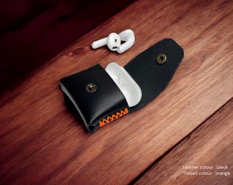 May include: Black leather AirPod case with orange stitching, open to reveal a white charging case. A pair of white wireless earbuds rests nearby on a wooden surface. The case has a snap closure.