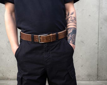 May include: A brown leather belt with a silver buckle. The belt is worn with black trousers and a black t-shirt. The belt has several holes for adjustment.