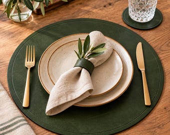 May include: A round dark green placemat featuring a beige speckled ceramic plate set, a beige linen napkin held by a dark green napkin ring, a gold fork, and a gold knife. The setting is arranged on a wooden surface with a glass.