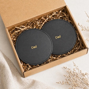 May include: Two black, round coasters, each with the word "Dad" in gold lettering, are presented in a cardboard gift box filled with brown paper. The coasters are made of a smooth material.