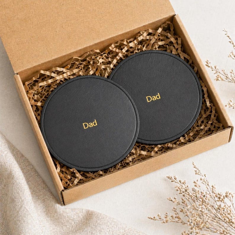 May include: Two black, round coasters, each with the word "Dad" in gold lettering, are presented in a cardboard gift box filled with brown paper. The coasters are made of a smooth material.