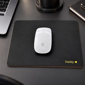 May include: A rectangular black mouse pad with rounded corners featuring the word Daddy and a small yellow heart icon printed in the bottom right corner. A white computer mouse with a silver logo is positioned in the centre of the mouse pad.