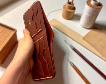 May include: Open, reddish-brown leather wallet with multiple card slots and white stitching. The wallet is held open, showcasing its interior. The word "ezzo" is visible on the wallet. Crafting tools, including needles and thread, are in the background.