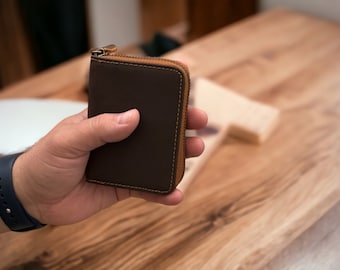 May include: A brown leather wallet with a zipper closure and a tan leather trim. The wallet is being held in a hand.