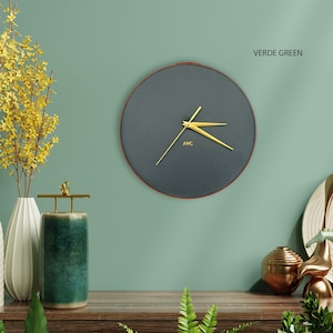 May include: A round, dark gray wall clock with gold hands and a gold rim. The clock face has the letters "AVC" printed in gold. The background is a light green wall with the text "VERDE GREEN".