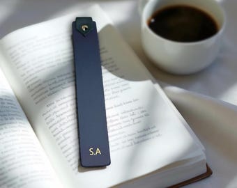May include: A navy blue leather bookmark with gold initials "S.A" rests in an open book. The bookmark features a dark green tab with a gold rivet. A white coffee cup filled with dark coffee is beside the book, suggesting a reading break.