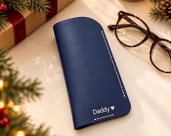 May include: A navy blue leather glasses case featuring white contrast stitching along the side and bottom edges. The front of the case is engraved with the word Daddy followed by a small heart symbol. The case has a smooth finish.