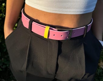 May include: A pink leather belt with a yellow stripe and a silver buckle. The belt is worn around the waist of a person wearing black trousers.
