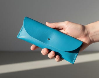 May include: A turquoise leather glasses case with a curved flap and a silver snap closure. The case is held in a hand, showcasing its smooth texture and vibrant colour. The case is designed to protect eyewear.