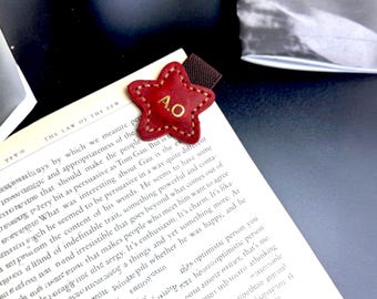 May include: A red leather star-shaped bookmark with gold stitching and the initials "A.O." in gold lettering. The bookmark is on top of a book with the text "The Law of the Few" visible.