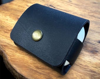 May include: A black leather case with a brass button fastening. The case is rectangular with curved edges and appears to be holding white objects. The case is on a wooden surface.