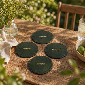 May include: Four dark green leather coasters, each with "Thompson" in gold lettering, sit on a wooden table. A vase of flowers, a glass of water, and a bowl of limes are also on the table.