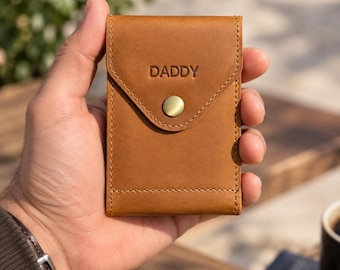May include: A tan leather card holder with a flap closure secured by a brass-colored metal snap button. The word DADDY is embossed in capital letters on the front flap. The wallet features visible stitching along the edges and a smooth finish.
