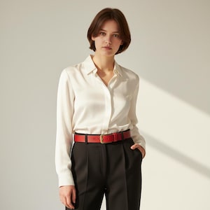 May include: A woman wearing a white button-down shirt, black pants, and a red leather belt with a gold-colored buckle. The belt is approximately 1.25 inches wide. The outfit is simple and classic.