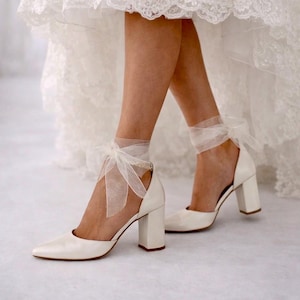 Op de afbeelding: Een paar ivoorkleurige satijnen pumps met blokhak, een puntige neus en een transparant organza lint dat om de enkel wordt gestrikt. De schoenen worden gedragen onder een witte kanten jurk met een asymmetrische zoom die de schoenen zichtbaar maakt.