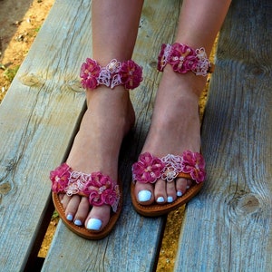 K&ouml;nnte beinhalten: Rosa und wei&szlig;e Blumensandalen mit Kn&ouml;chelriemen. Die Sandalen haben eine Ledersohle und sind aus Stoff.