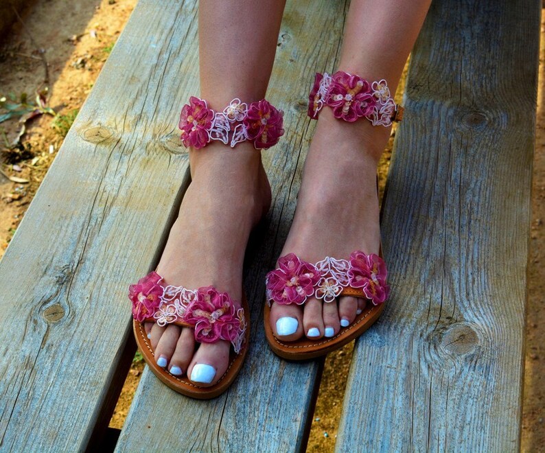 K&ouml;nnte beinhalten: Rosa und wei&szlig;e Blumensandalen mit Kn&ouml;chelriemen. Die Sandalen haben eine Ledersohle und sind aus Stoff.