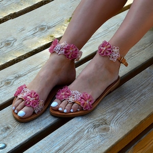 K&ouml;nnte beinhalten: Ein Paar braune Ledersandalen mit rosa und wei&szlig;en Blumenverzierungen. Die Sandalen haben einen Riemen, der um den Kn&ouml;chel geht, und einen Schnallenverschluss.