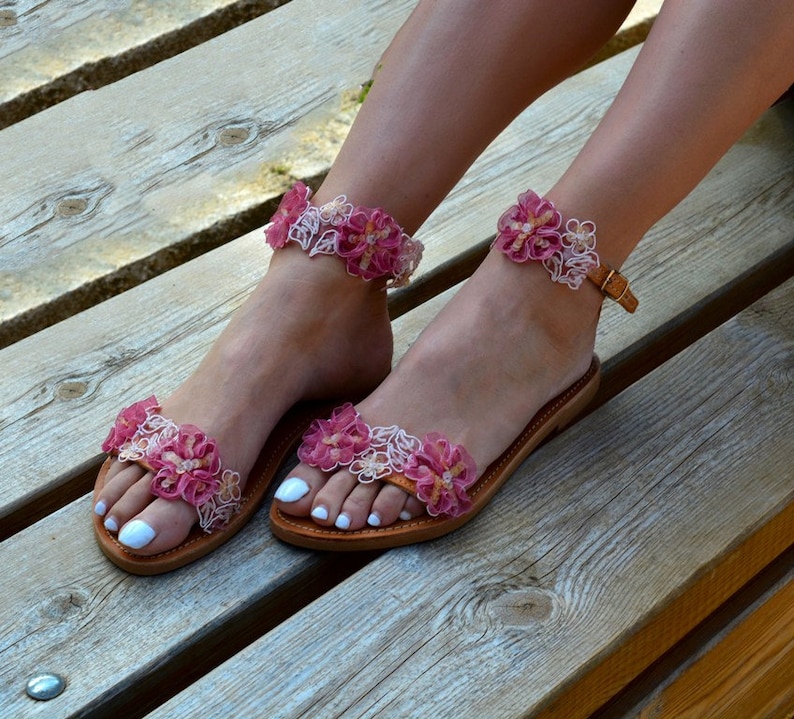 K&ouml;nnte beinhalten: Ein Paar braune Ledersandalen mit rosa und wei&szlig;en Blumenverzierungen. Die Sandalen haben einen Riemen, der um den Kn&ouml;chel geht, und einen Schnallenverschluss.