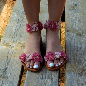 K&ouml;nnte beinhalten: Ein Paar flache Sandalen mit rosa und wei&szlig;en Blumen. Die Sandalen haben eine zarte Spitze und sind perfekt f&uuml;r den Sommer.