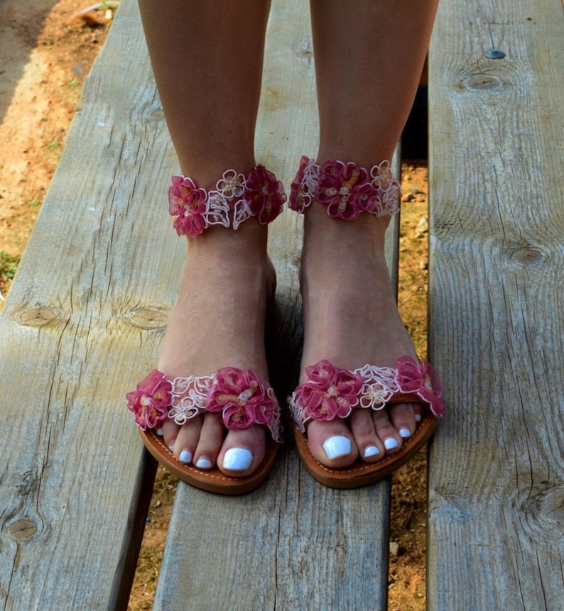 K&ouml;nnte beinhalten: Ein Paar flache Sandalen mit rosa und wei&szlig;en Blumen. Die Sandalen haben eine zarte Spitze und sind perfekt f&uuml;r den Sommer.