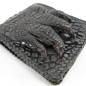 May include: A dark brown square wallet featuring a textured, embossed surface that mimics the appearance of reptile scales. The front of the wallet displays a prominent, three-dimensional sculpted claw design with sharp, curved talons.