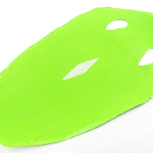 Puede incluir: Una piel de raya de color verde lima brillante con una superficie texturizada y granulada, que presenta un parche dorsal calcificado blanco en forma de diamante alargado en el centro y dos pequeños cortes simétricos cerca del borde superior.