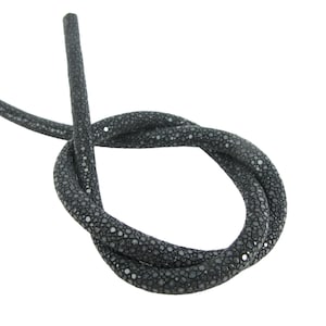Puede incluir: Un cordón cilíndrico y flexible con una superficie de color gris carbón oscuro y una textura granulada y distintiva. El cordón está atado en un nudo simple y suelto, mostrando el patrón circular y uniforme del material.