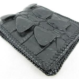 May include: A black bi-fold wallet featuring a textured, raised scale pattern on the exterior surface. The edges of the wallet are finished with a detailed, braided leather trim that runs along the entire perimeter of the folded accessory.